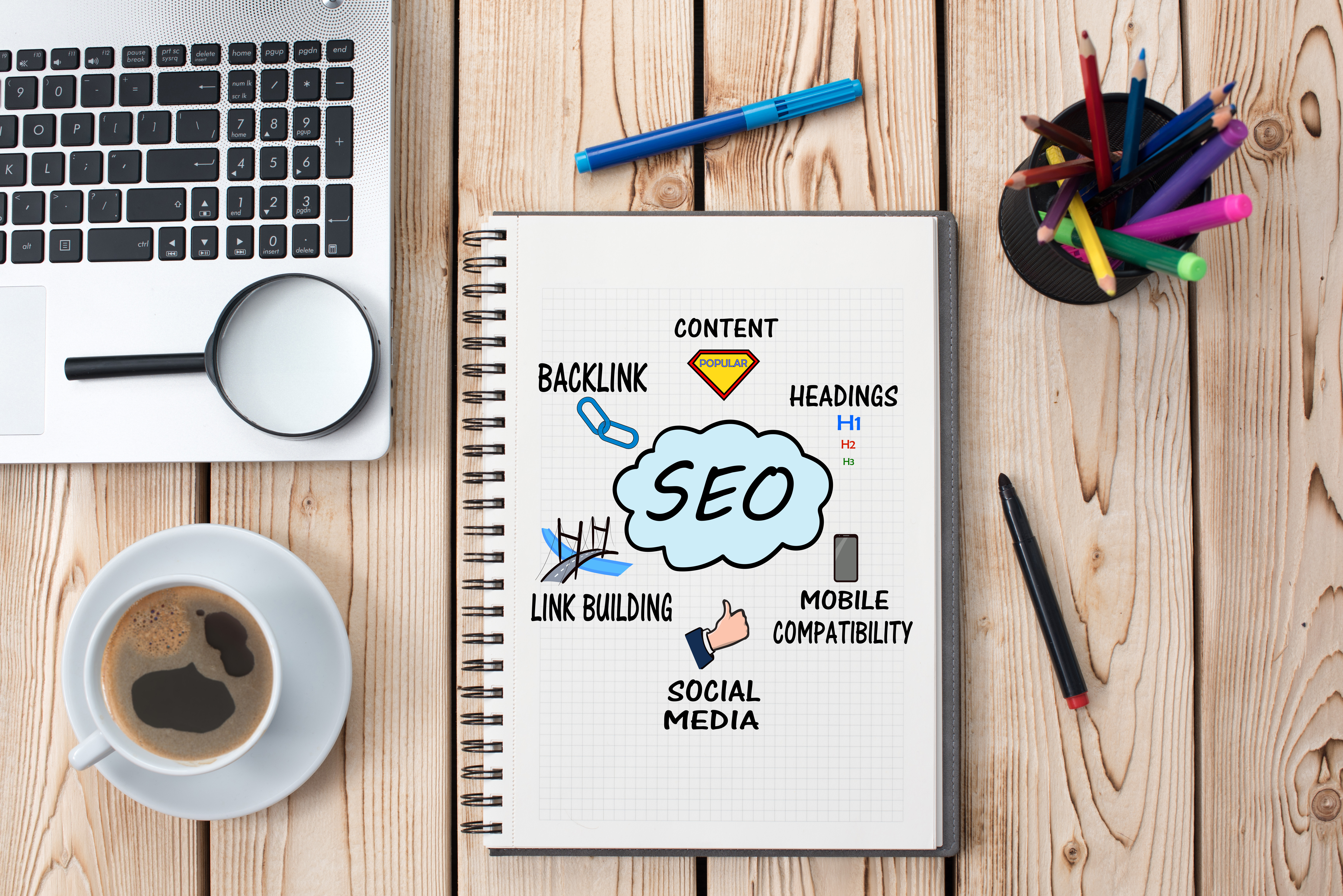 Image depicting a working laptop, coffee, pens, and a notebook which appears to store notes on Search Engine Optimization.